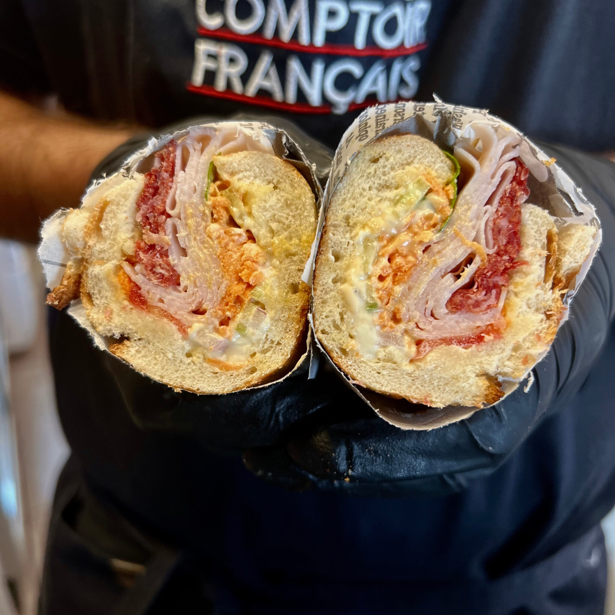 Sandwich charcuterie coupé en deux : salami français, dinde et crudités, tenu devant le tablier Comptoir Français