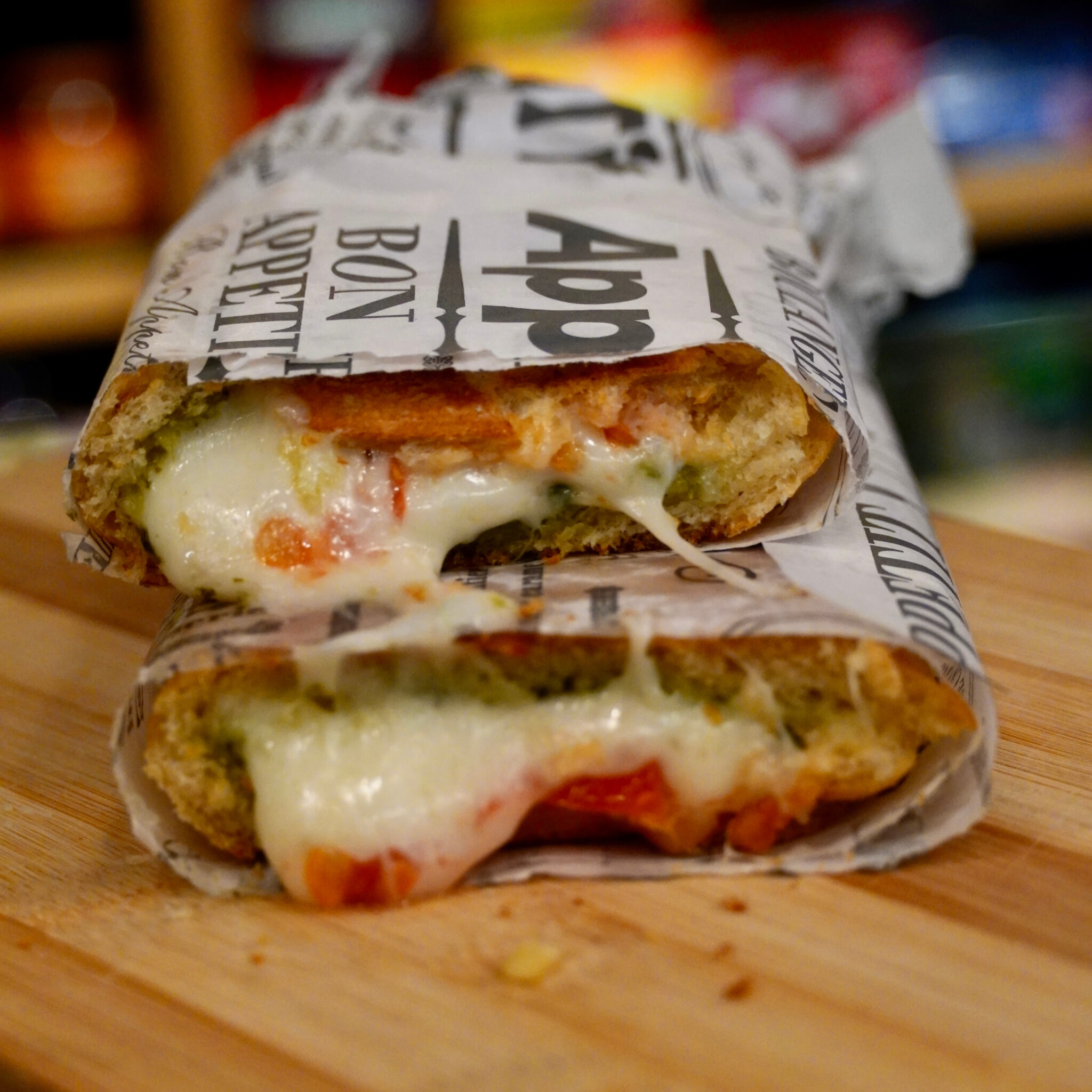 Panini mozzarella pesto ouvert en deux sur une planche en bois, dans son papier Bon Appétit